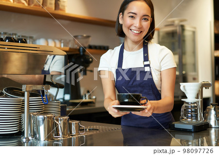 Portrait of smiling young woman providing customer service in cafe, holding cup of coffee, giving order to client, wearing apron uniform Portrait of smiling young woman providing customer service in cafe, holding cup of coffee, giving order to client, wearing apron uniform 98097726