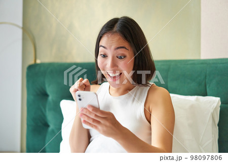 Winning and triumphing. Excited girl in bed, looking at her phone with amazed, surprised face, celebrating victory, reading good news Winning and triumphing. Excited girl in bed, looking at her phone with amazed, surprised face, celebrating victory, reading good news 98097806