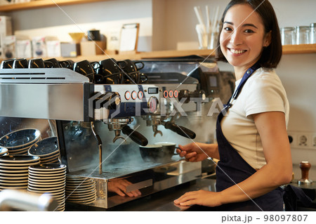 Smiling cute korean girl, barista working with coffee machine, prepare cappuccino, using steamr at cafe counter Smiling cute korean girl, barista working with coffee machine, prepare cappuccino, using steamr at cafe counter 98097807