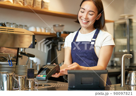 Smiling asian girl barista, giving client POS terminal, help pay with credit card, standing at counter in coffee shop, working in cafe 98097821