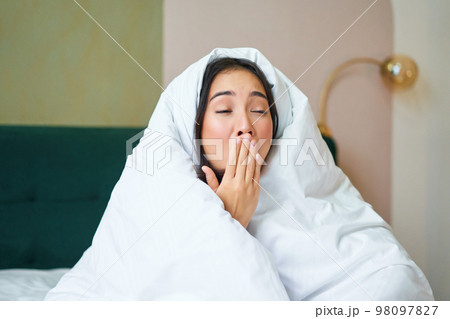 Close up of funny sleepy girl, asian woman yawns after waking up early morning, covers herself with blanket duvet, holds hand near opened mouth Close up of funny sleepy girl, asian woman yawns after waking up early morning, covers herself with blanket duvet, holds hand near opened mouth 98097827