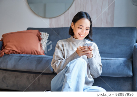 People and technology. Smiling asian woman sitting at home, using her mobile phone, typing message, browing internet or shopping online from smartphone app 98097956