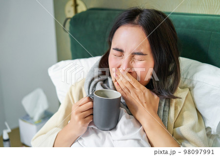 Close up of asian girl in bed, feeling sick, catching cold and sneezing, wiping runny nose, drinking hot tea 98097991