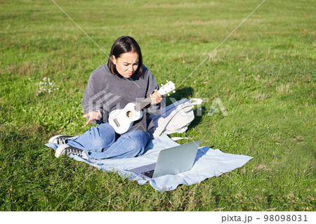 Cute asian girl, musician studying, looks at tutorial on her laptop, online tutor shows how to play ukulele, student sits on grass in park on sunny day Cute asian girl, musician studying, looks at tutorial on her laptop, online tutor shows how to play ukulele, student sits on grass in park on sunny day 98098031