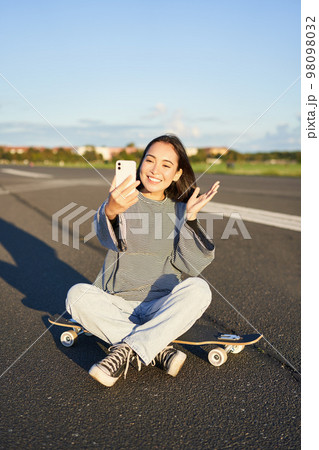 Happy teen asian girl takes selfie, video chats on smartphone app while sits on skateboard, skater records vlog while sits on longboard 98098032