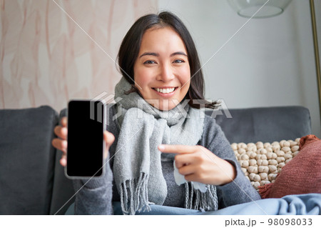 Portrait of smiling girl staying ill at home, showing mobile phone screen, recommending online medical application, pharmacy or gp on smartphone 98098033