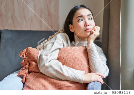 Women and wellbeing concept. Portrait of thoughtful asian woman sitting on sofa with pillow, looking aside with thinking concentrated face 98098118