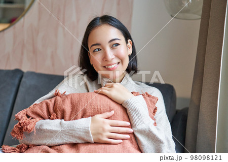 Women and wellbeing concept. Young woman in warm clothes, sits at home, hugs pillow on sofa and smiles pleased, thinks of smth 98098121