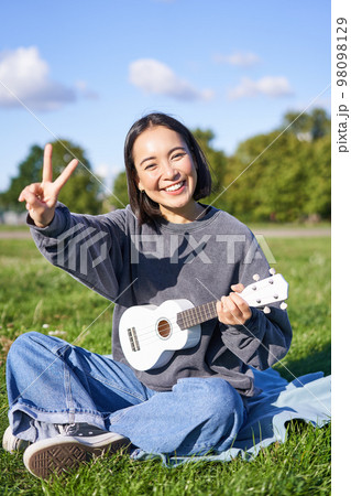 Vertical shot of positive asian girl shows peace sign, plays ukulele in park, rests and enjoys the day. Copy space Vertical shot of positive asian girl shows peace sign, plays ukulele in park, rests and enjoys the day. Copy space 98098129