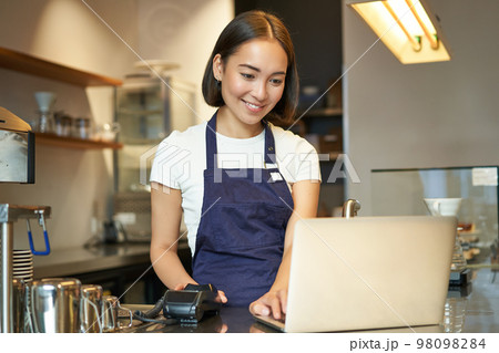 Portrait of young girl cafe owner, looking at her laptop, taking order, serving customer in coffee shop 98098284