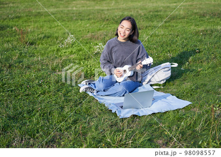 Cute korean girl, musician sits in park, plays ukulele and sings, looks up chords and tutorials on laptop 98098557