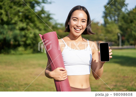 Smiling asian fitness girl with rubber yoga mat, shows her smartphone screen, recommends workout application, stands on lawn in park Smiling asian fitness girl with rubber yoga mat, shows her smartphone screen, recommends workout application, stands on lawn in park 98098608