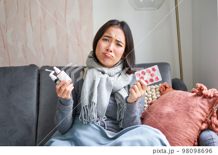 Korean woman showing medical pills, sore throat sprays, feeling sick, staying at home as feeling unwell, catching cold in winter Korean woman showing medical pills, sore throat sprays, feeling sick, staying at home as feeling unwell, catching cold in winter 98098696