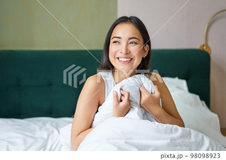 Close up of happy beautiful asian woman, waking up in bed and enjoys morning, looking outside window with sleepy smile on her face, hugging duvet 98098923