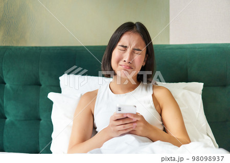 Disappointed asian girl, crying and whining, feeling upset, lying in bed on pillow, holding mobile phone, read upsetting bad news, waking up in awful mood Disappointed asian girl, crying and whining, feeling upset, lying in bed on pillow, holding mobile phone, read upsetting bad news, waking up in awful mood 98098937