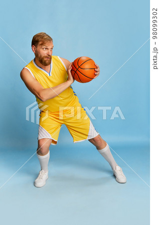Happy man with beard wearing sportswear doing exercises with basketball ball isolated over blue background. Concept of active lifestyle, positive emotions, sport 98099302