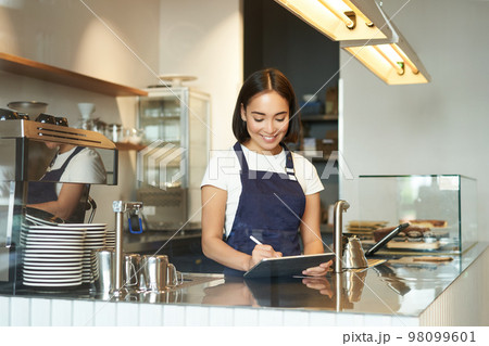 Smiling cute barista girl, korean woman working in cafe, using tablet to process order, making coffee at counter 98099601