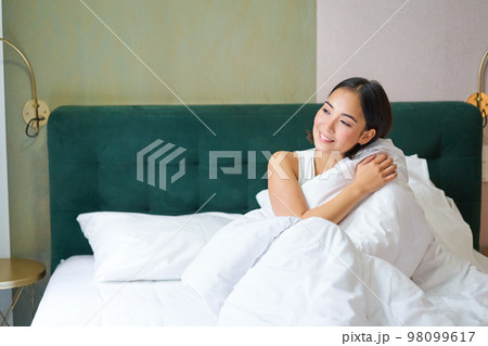 Beautiful sleepy asian girl wakes up in her bed with cozy white blankets, looking outside window with pleased smile. Lifestyle concept 98099617