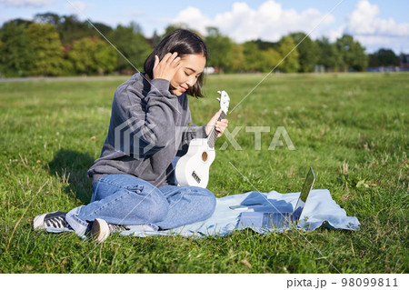 Smiling asian girl learns how to play ukulele via laptop, online video tutorials, sitting on grass in park with musical instrument Smiling asian girl learns how to play ukulele via laptop, online video tutorials, sitting on grass in park with musical instrument 98099811
