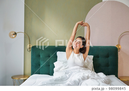 Beautiful asian woman lying in her bedroom on bed, stretching hands and looking outside, waking up from sleep Beautiful asian woman lying in her bedroom on bed, stretching hands and looking outside, waking up from sleep 98099845