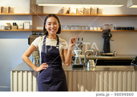 Portrait of asian girl in apron, standing in cafe near counter, barista pointing finger at copy space, empty banner coffee shop Portrait of asian girl in apron, standing in cafe near counter, barista pointing finger at copy space, empty banner coffee shop 98099860