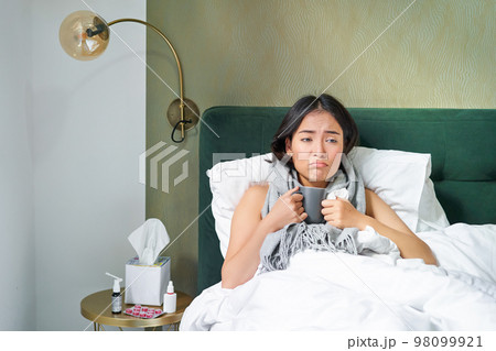 Sick sad asian girl lying in her bed with flu, catch a cold, taking pills and drinking hot tea, feeling upset Sick sad asian girl lying in her bed with flu, catch a cold, taking pills and drinking hot tea, feeling upset 98099921