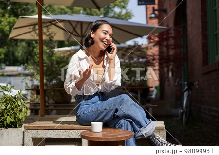 Beautiful young asian woman sitting in cafe with cup of coffee, talking on mobile phone, having lively conversation Beautiful young asian woman sitting in cafe with cup of coffee, talking on mobile phone, having lively conversation 98099931
