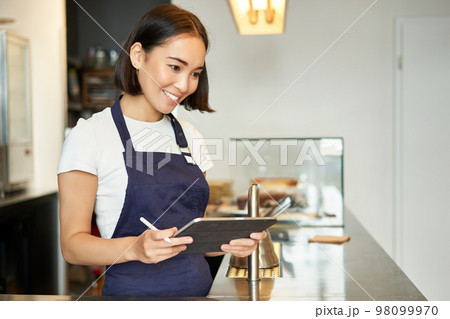 Small cafe business. Smiling asian girl barista in apron, using tablet as POS terminal, processing coffee order behind counter Small cafe business. Smiling asian girl barista in apron, using tablet as POS terminal, processing coffee order behind counter 98099970