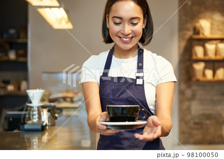 Smiling korean girl in uniform, working in cafe, giving you cup of coffee, preparing order for client 98100050