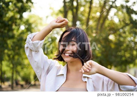 People lifestyle. Portrait of young brunette woman dancing, smiling and laughing, walking in park with hands lift up high, enjoying summer day outside 98100254