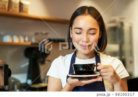 Close up of smiling asian girl barista, worker in cafe, drinking coffee on her break, holding cup with pleased face and closed eyes, enjoying her drink 98100317