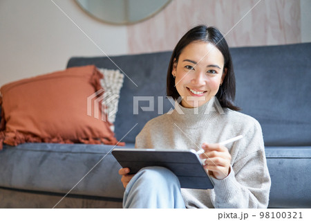Portrait of asian woman with tablet, drawing, working on design project, holding pen, sitting in her living room, freelancing Portrait of asian woman with tablet, drawing, working on design project, holding pen, sitting in her living room, freelancing 98100321