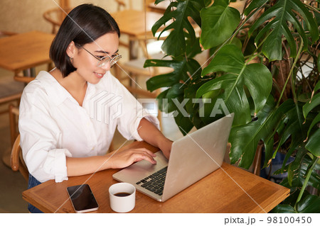 Working female entrepreneur sitting in cafe with laptop, typing on computer, drinking coffee 98100450