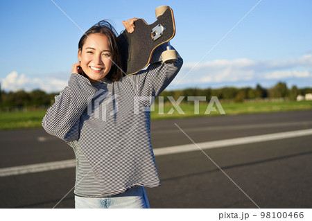Portrait of beautiful young skater girl, standing with longboard and smiling at camera. Asian woman with skateboard standing on road Portrait of beautiful young skater girl, standing with longboard and smiling at camera. Asian woman with skateboard standing on road 98100466