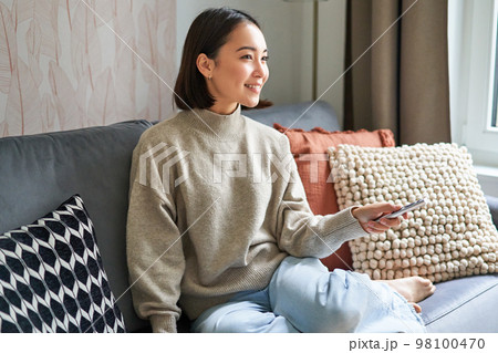 Beautiful asian girl in cozy home, watching television, holding remote and switching channels, sitting on sofa 98100470