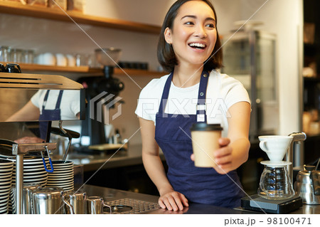 Smiling asian barista girl, giving takeaway coffee cup, prepare takeout order to guest in cafe, wearing apron uniform 98100471