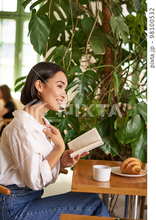 Vertical shot of asian brunette girl sitting elegant with book, reading in cafe and smiling, drinking coffee Vertical shot of asian brunette girl sitting elegant with book, reading in cafe and smiling, drinking coffee 98100532