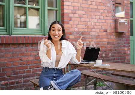 Stylish urban girl, asian woman with laptop sits in cafe, points at upper right corner banner, shows copy space with surprised face expression Stylish urban girl, asian woman with laptop sits in cafe, points at upper right corner banner, shows copy space with surprised face expression 98100580