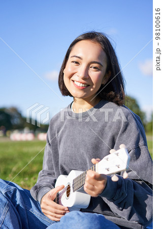 Portrait of cute smiling girl playing ukulele in park. Young woman with musical instrument sitting outdoors 98100616