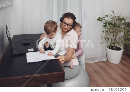 Mother sits on a fitness ball in headphones and works at home with her children during quarantine Mother sits on a fitness ball in headphones and works at home with her children during quarantine 98101205