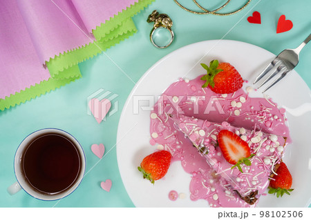 A slice of cheesecake decorated with strawberries, pink frosting on a white plate. Valentine's Day dessert. Hot strawberry drink in a cup, ring with rabbit - symbol of the year, hearts, top view. A slice of cheesecake decorated with strawberries, pink frosting on a white plate. Valentine's Day dessert. Hot strawberry drink in a cup, ring with rabbit - symbol of the year, hearts, top view. 98102506