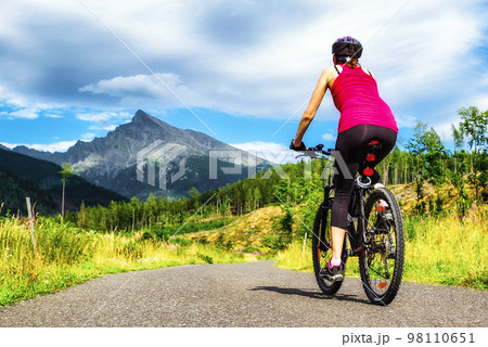Woman riding mountain bike on road in forest with hill at background in High Tatras mountains, Slovakia Woman riding mountain bike on road in forest with hill at background in High Tatras mountains, Slovakia 98110651