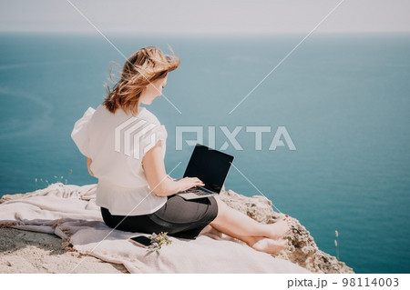 Woman sea laptop. Successful business woman working on laptop by the sea. Pretty lady typing on computer at summer day outdoors. Freelance, digital nomad, travel and holidays concept. 98114003