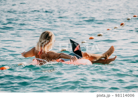 Woman works on laptop in sea. Freelancer, young blond woman in sunglases floating on an inflatable big pink donut with a laptop in the sea at sunset. Freelance, travel and holidays concept. 98114006