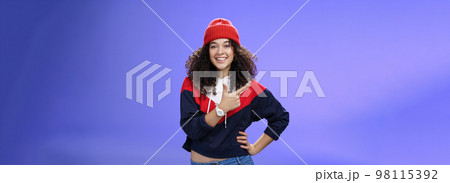 Studio shot of happy charming stylish city girl in beanie and warm sweatshirt ready rock winter up pointing at upper left corner and smiling delighted showing cool place over blue wall Studio shot of happy charming stylish city girl in beanie and warm sweatshirt ready rock winter up pointing at upper left corner and smiling delighted showing cool place over blue wall 98115392