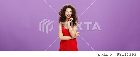 Gorgeous lady in red feeling shy and awkward being invited for dance standing timid and feminine over purple background touching strand of curly hair smiling broadly and crossing arm over chest 98115393