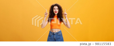 Portrait of confident charismatic caucasian female with curly hairstyle in red lipstick and orange cropped top pointing up with raised hands and smiling cheerfully at camera assured copy space fine 98115583