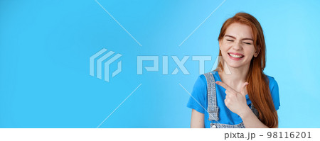 Cheerful carefree redhead woman sincere smile, pointing left introduce copy space joyfully close eyes, show white toothy grin delighted, feel upbeat rejoicing, joyful positive mood, blue background 98116201