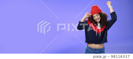 Girl celebrating favorite season in year. Portrait of carefree and joyful dancing woman with curly hair in cute red hat lifting hands in dance movements smiling enjoying music and holidays 98116337