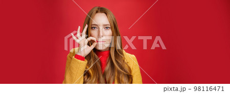 Girl putting seal on mouth making promise not tell anyone secret, sucking lips and holding finger near as zipping not spill words, looking serious and determined to keep surprise safe over red wall 98116471
